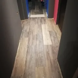 Korytarz z nową podłogą z paneli imitujących drewno, ściany w odcieniach szarości, błękitu i różu, widok z perspektywy wejścia, z widocznym fragmentem pomieszczenia w oddali.