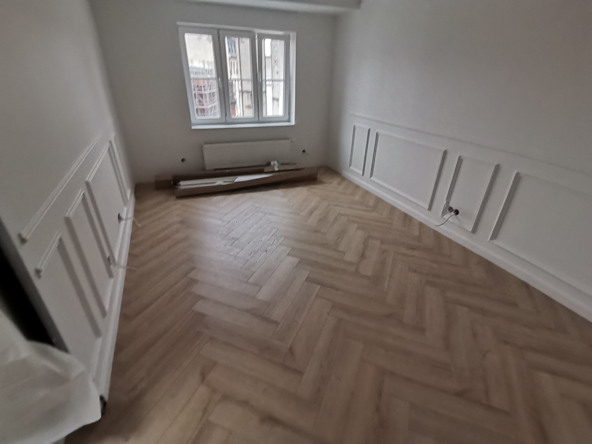 Wnętrze pokoju po remoncie z nową podłogą ułożoną w jodełkę i dekoracyjnymi panelami ściennymi, widoczne kartony z materiałami budowlanymi pod oknem.