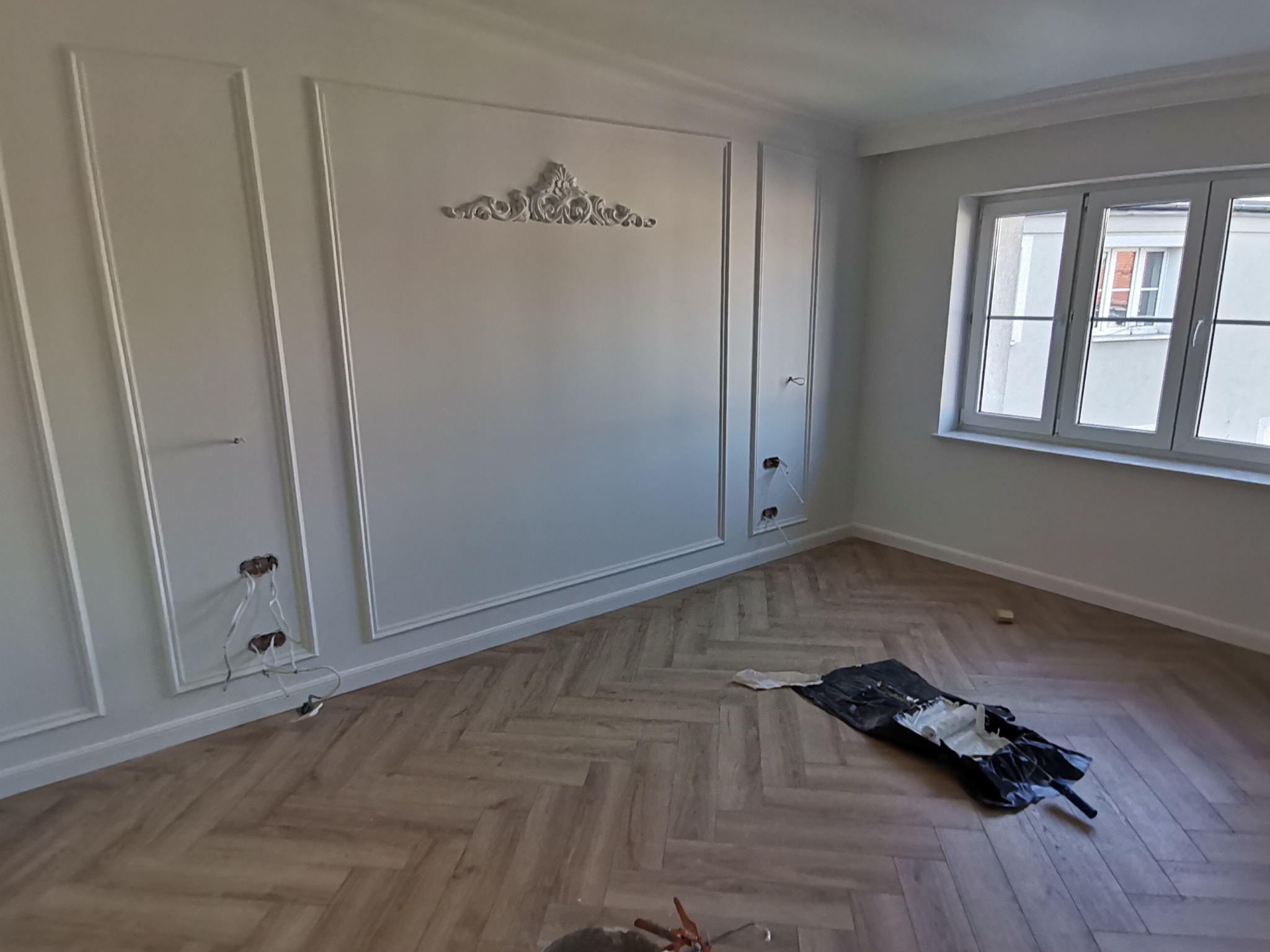 Wykończone wnętrze pokoju z dekoracyjnymi listwami ściennymi, rozetą nad listwami i drewnianą podłogą ułożoną w jodełkę. Widoczne wystające kable elektryczne ze ściany oraz resztki materiałów...