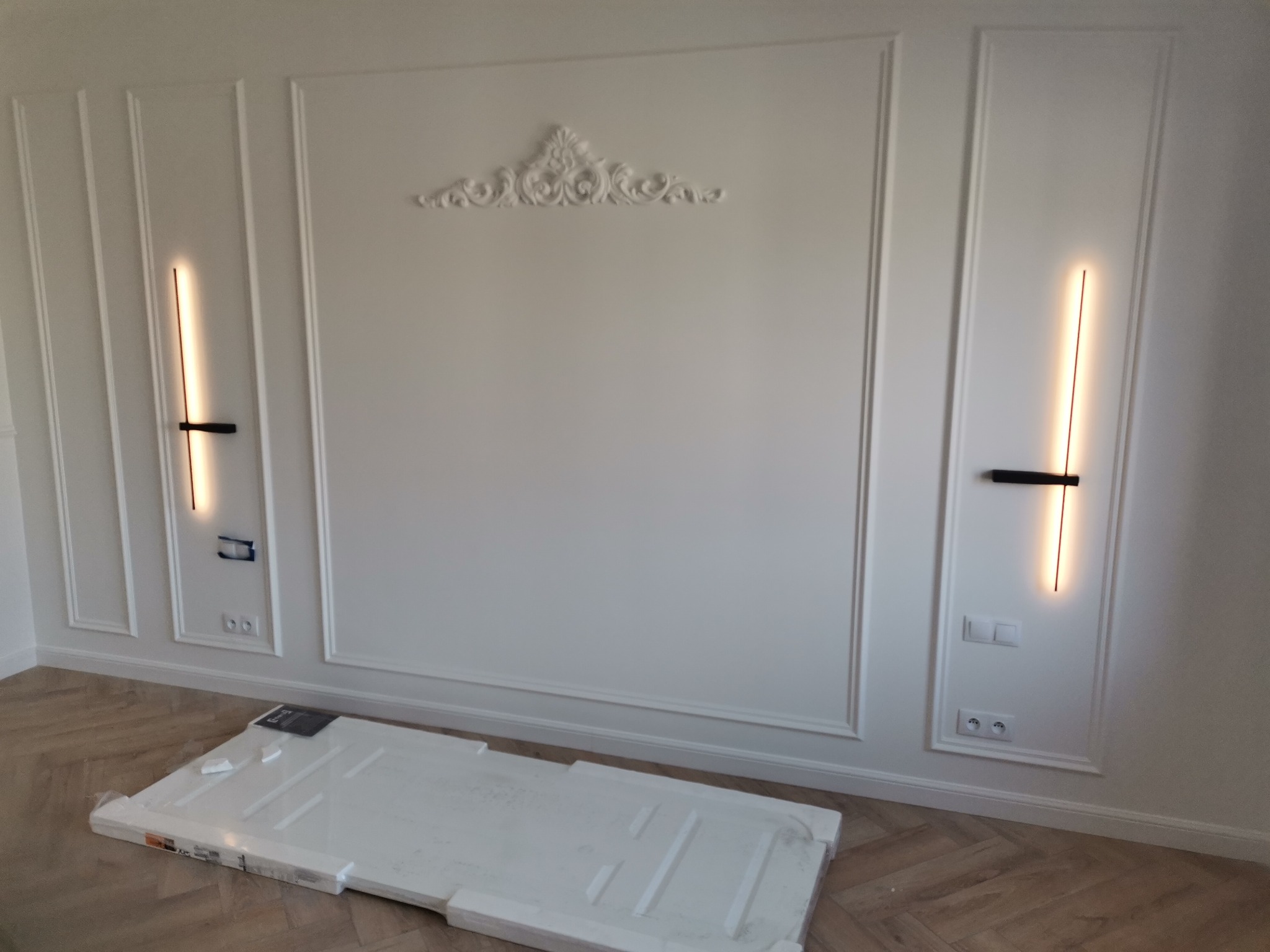 Biała ściana z dekoracyjną sztukaterią, nowoczesnymi kinkietami i zapakowanymi elementami wykończeniowymi na podłodze z parkietu ułożonego we wzór jodełki.