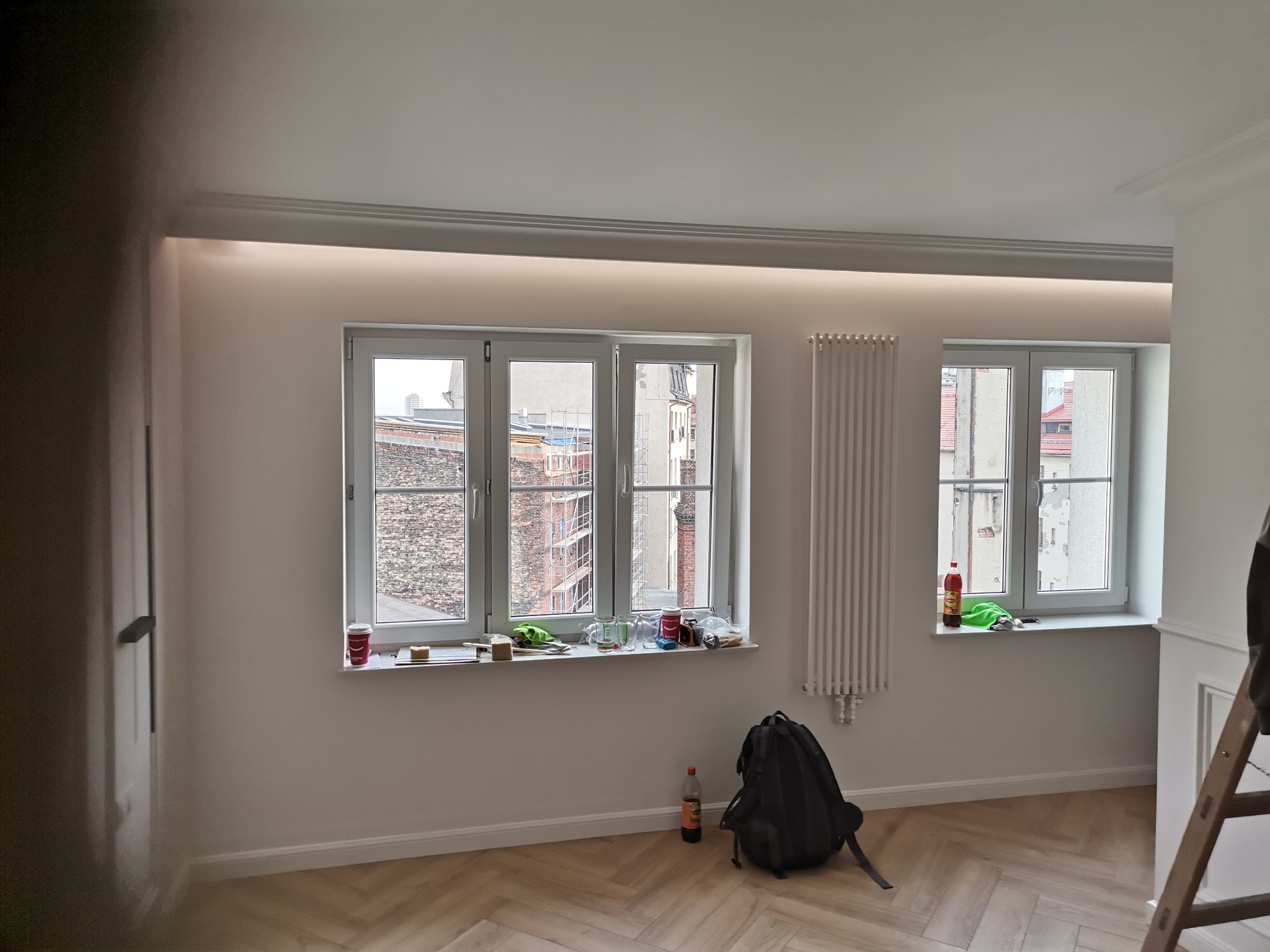 Pokój po remoncie z dwiema oknami, grzejnikiem, plecakiem, butelką i schodami, parkiet ułożony w jodełkę, sztukateria sufitowa z oświetleniem LED.