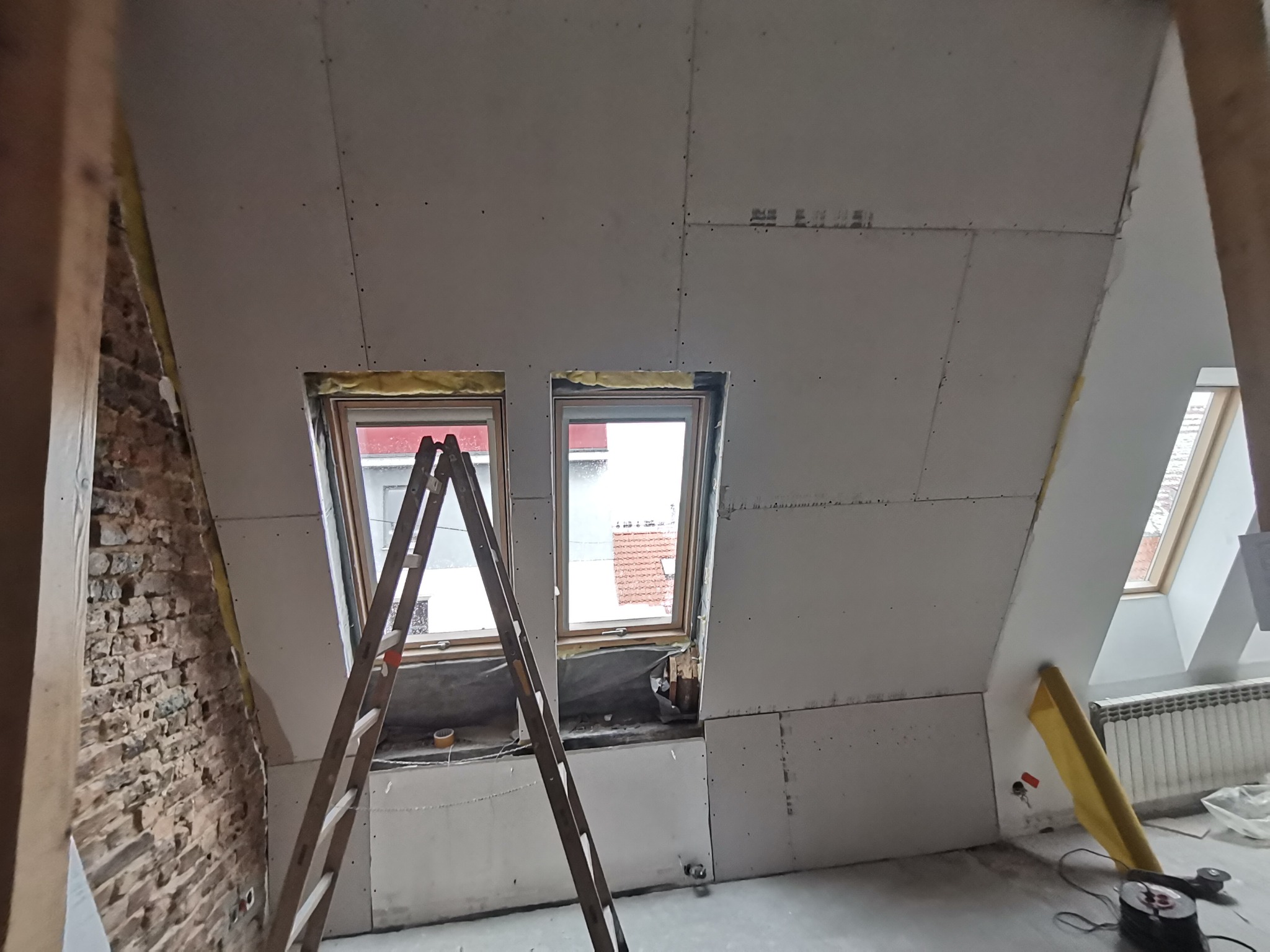 Poddasze w trakcie wykańczania płytami gipsowo-kartonowymi, widoczne dwie drewniane drabiny opierające się o okna dachowe, surowa ściana z cegły z boku i fragment grzejnika pod oknem po prawej.