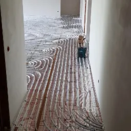 Wnętrze w trakcie montażu ogrzewania podłogowego: białe rurki ułożone na folii z czerwoną siatką, widoczne surowe ściany i drewniany szkielet sufitu, narzędzia Makity w tle.