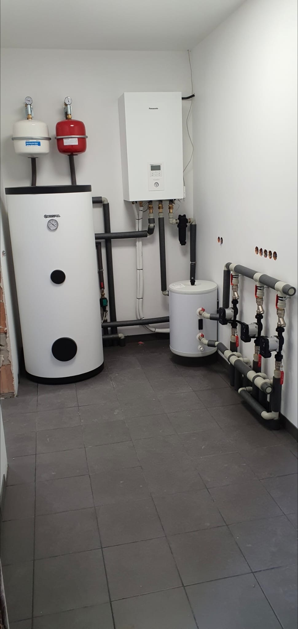 Biały bojler Kospel z manometrem i czarnymi okrągłymi otworami rewizyjnymi, obok biały zbiornik wyrównawczy i czerwony zbiornik przeponowy oraz wiszący piec gazowy Panasonic, połączone rurami...