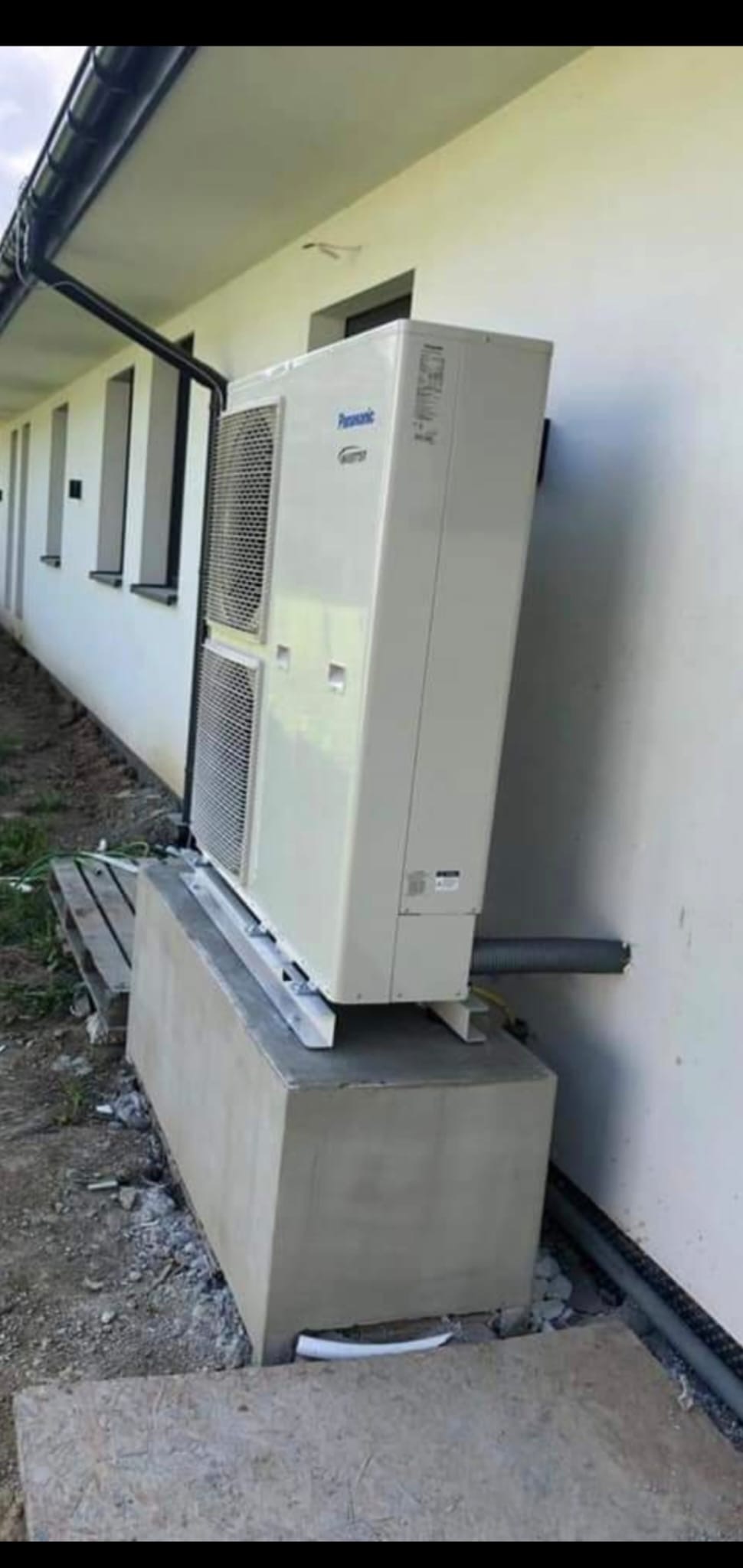Biała pompa ciepła Panasonic zamontowana na betonowym fundamencie przy ścianie budynku z widocznymi rurami i oknami