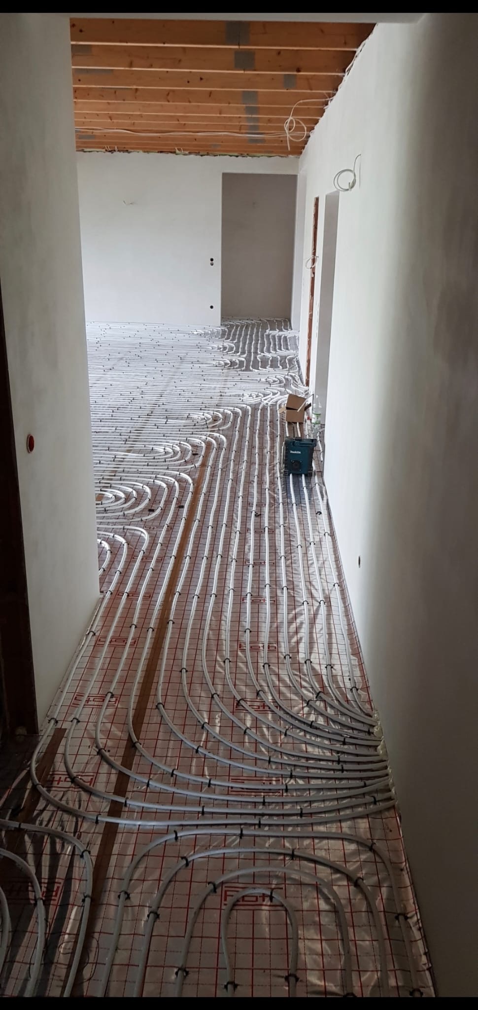 Wnętrze w trakcie montażu ogrzewania podłogowego: białe rurki ułożone na folii z czerwoną siatką, widoczne surowe ściany i drewniany szkielet sufitu, narzędzia Makity w tle.