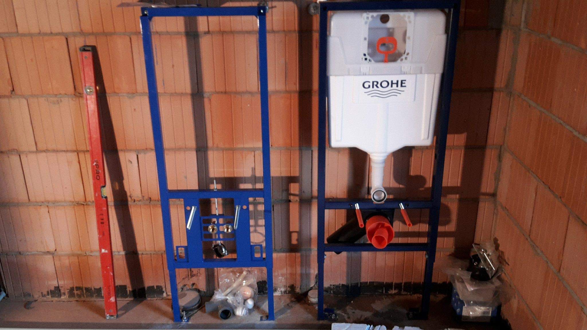 Stelaż podtynkowy WC marki Grohe zamontowany na tle ściany z czerwonej cegły, obok pionowa poziomica i elementy instalacji w folii.