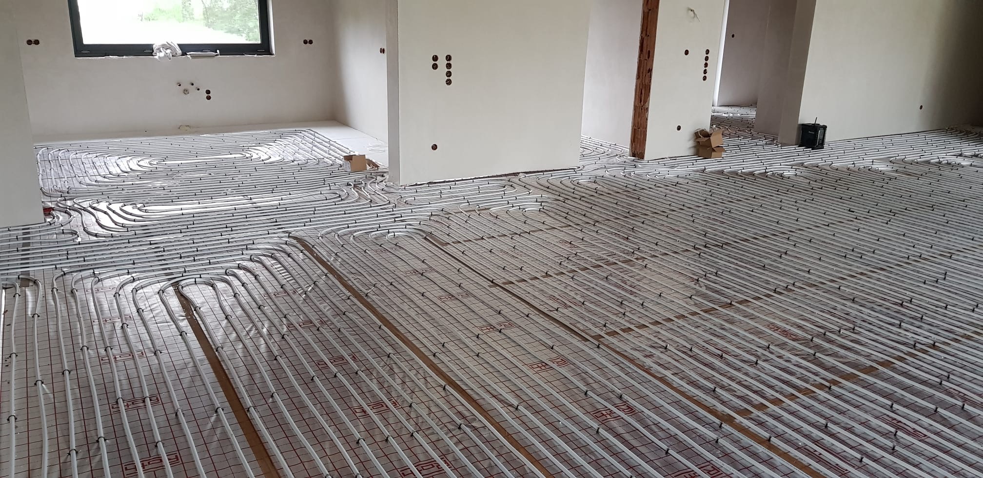 Rozległa instalacja ogrzewania podłogowego w surowym stanie, rury grzewcze rozłożone na siatce z folią izolacyjną, widoczne wystające kable elektryczne i puszki instalacyjne w ścianach...