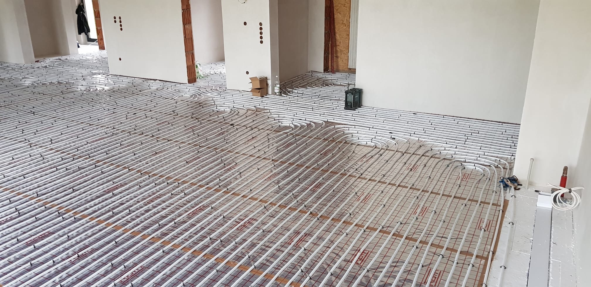 Rozłożona instalacja ogrzewania podłogowego na siatce montażowej, widoczne białe rurki i czerwona siatka, w tle surowe ściany i przygotowane podłoże.