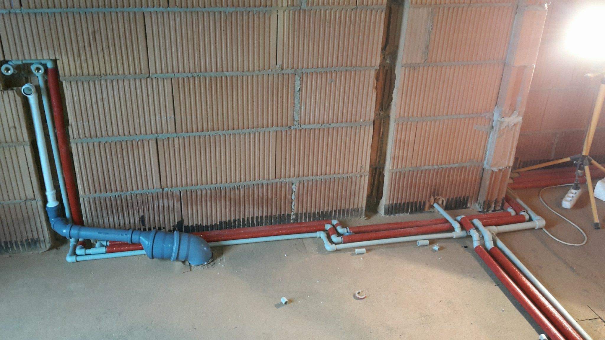 Surowy stan: Instalacja wodno-kanalizacyjna w trakcie montażu w budynku z cegły dziurawki, widoczne rury czerwone i szare, lampa budowlana na trójnogu w tle.