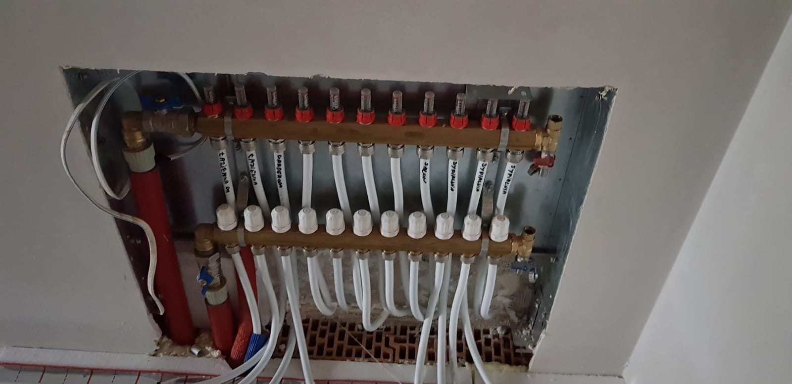 Rozdzielacz ogrzewania podłogowego z rurami oznaczonymi nazwami pomieszczeń, zamontowany w płycie gipsowo-kartonowej, widok od dołu.