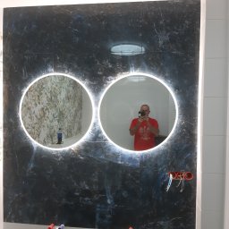 Specbud - Łazienka z dwoma okrągłymi lustrami z podświetleniem LED na ścianie z płytek imitujących ciemny marmur. Widoczne niezakończone podłączenia elektryczne i hydrauliczne.