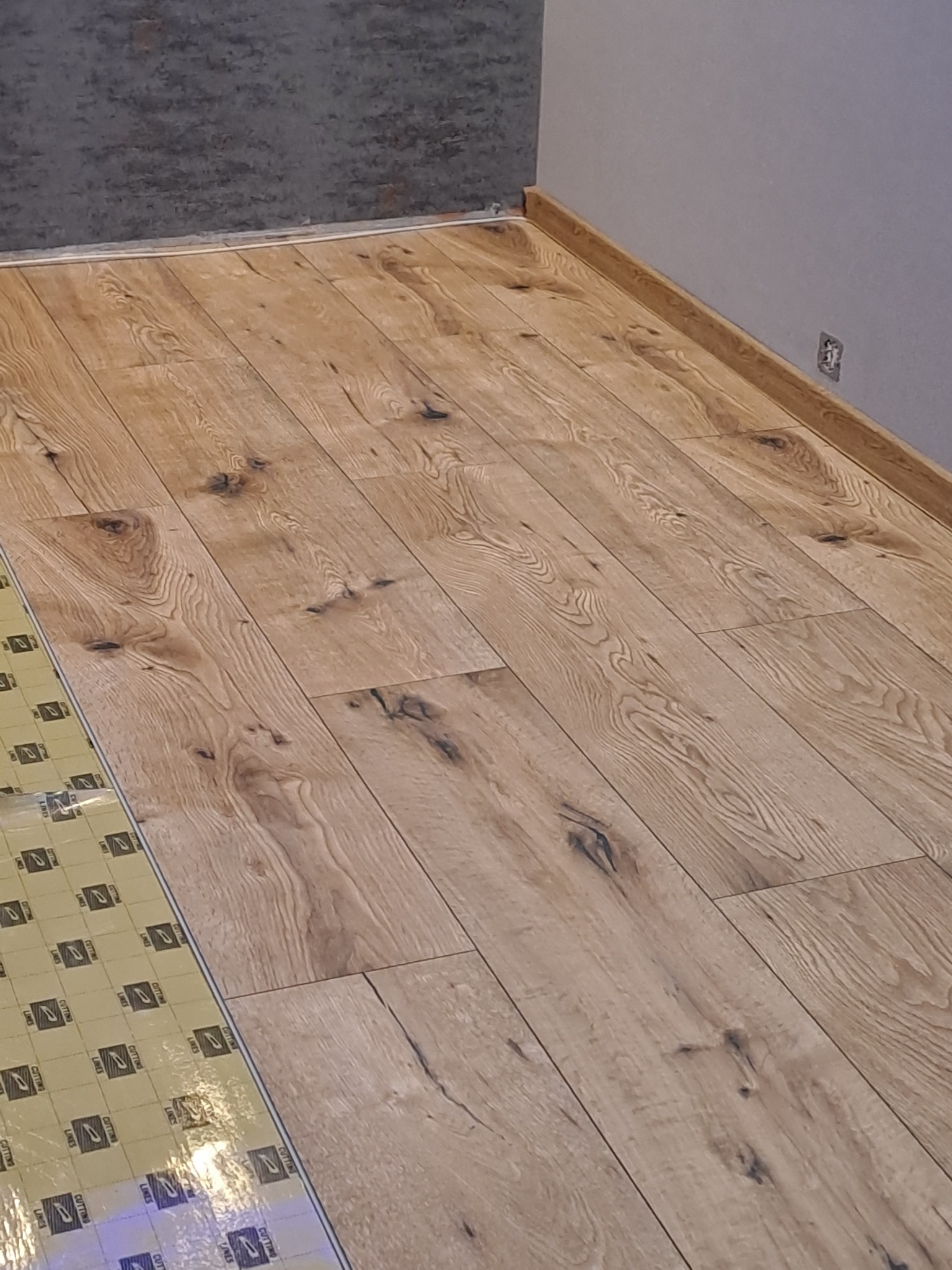 Układanie podłogi z drewnianych paneli, widoczny fragment podkładu izolacyjnego, ściana szara i wykończenie przy podłodze.