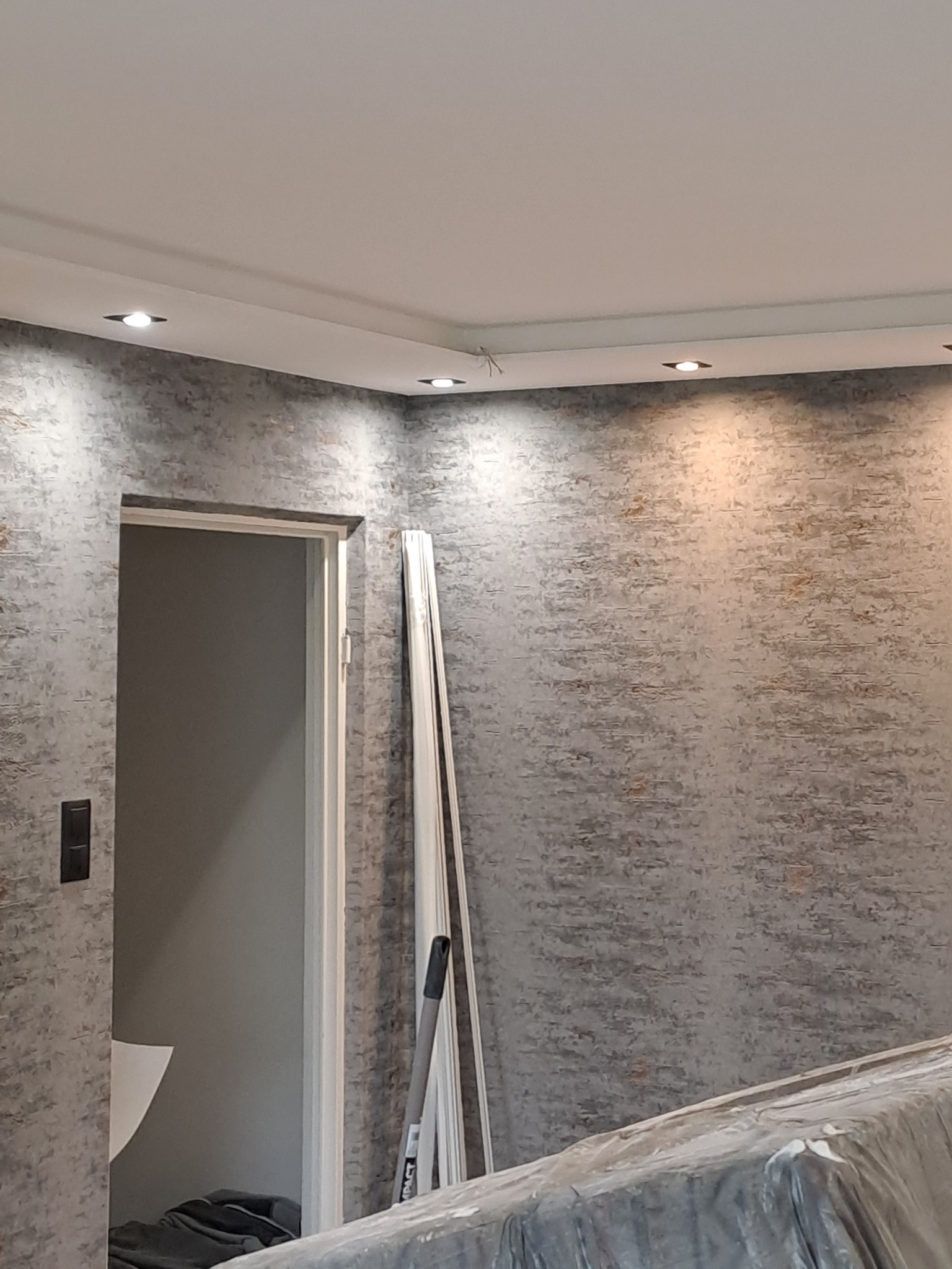 Ściana z dekoracyjną szarą fakturą, oświetlenie punktowe w suficie, biała ościeżnica drzwiowa, listwy przypodłogowe oparte o ścianę, zabezpieczone folią meble w pomieszczeniu w trakcie prac.