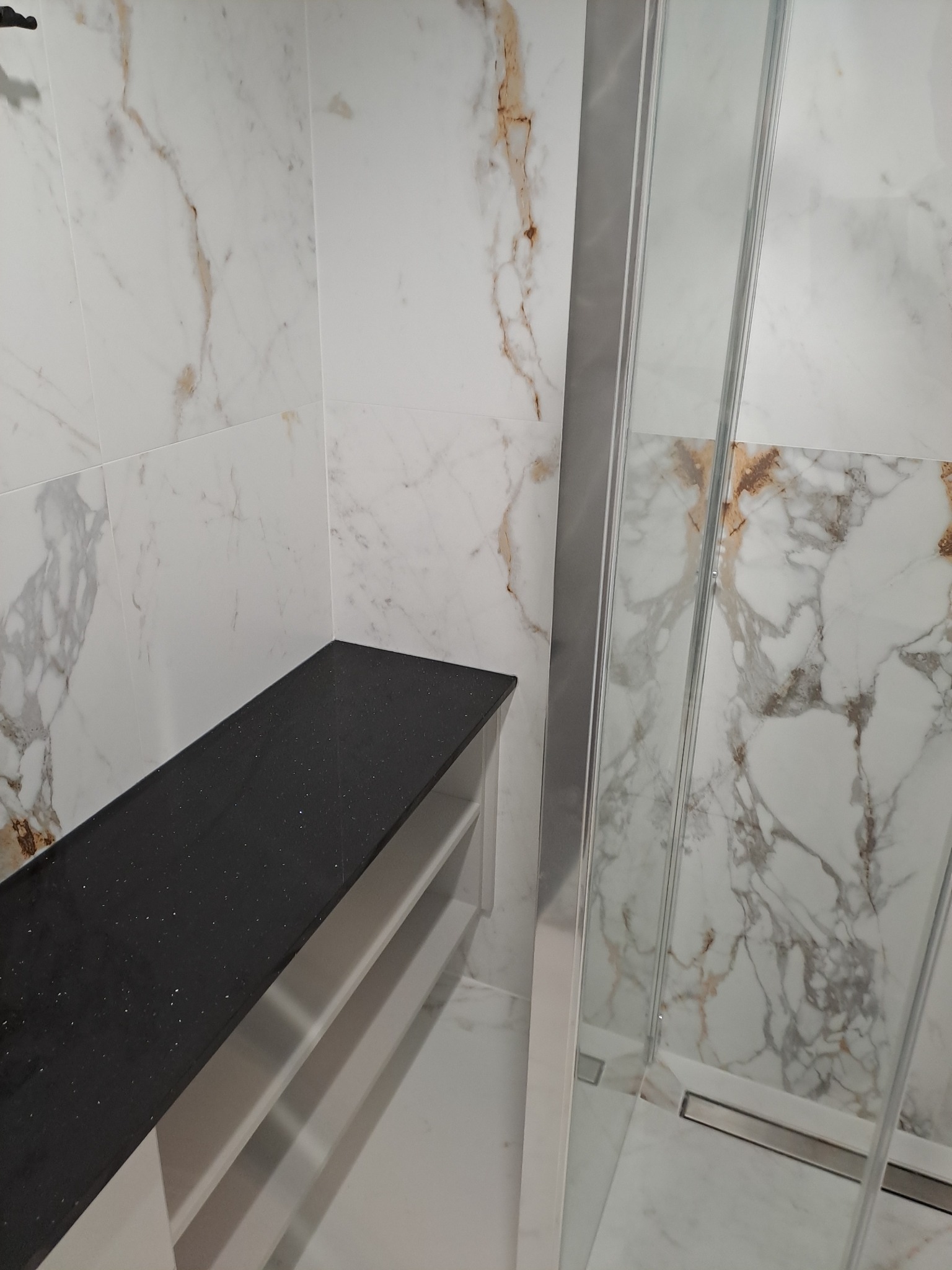 Wykończona łazienka z marmurowymi płytkami, czarnym blatem z brokatem i szklaną kabiną prysznicową z odpływem liniowym w podłodze.