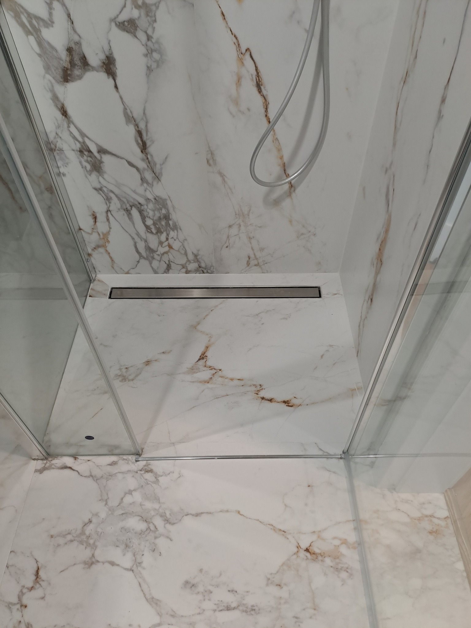 Kabina prysznicowa walk-in z marmurowymi płytami, odpływ liniowy w posadzce, chromowana słuchawka prysznicowa, minimalistyczny design.