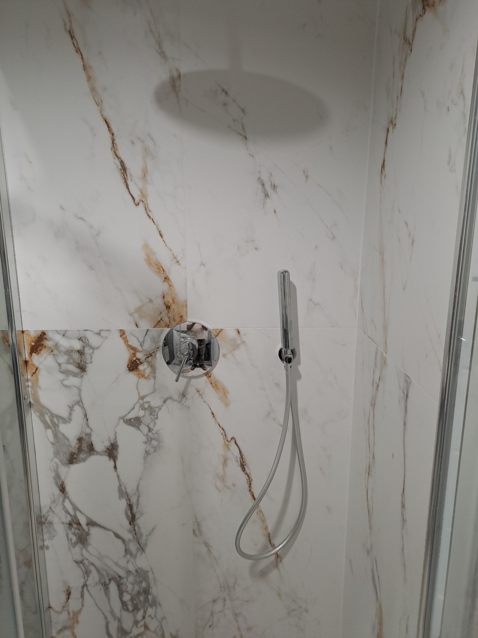 Kabina prysznicowa wykończona białymi płytkami imitującymi marmur z brązowymi żyłami, chromowana bateria z mieszaczem i słuchawka prysznicowa na wężu.