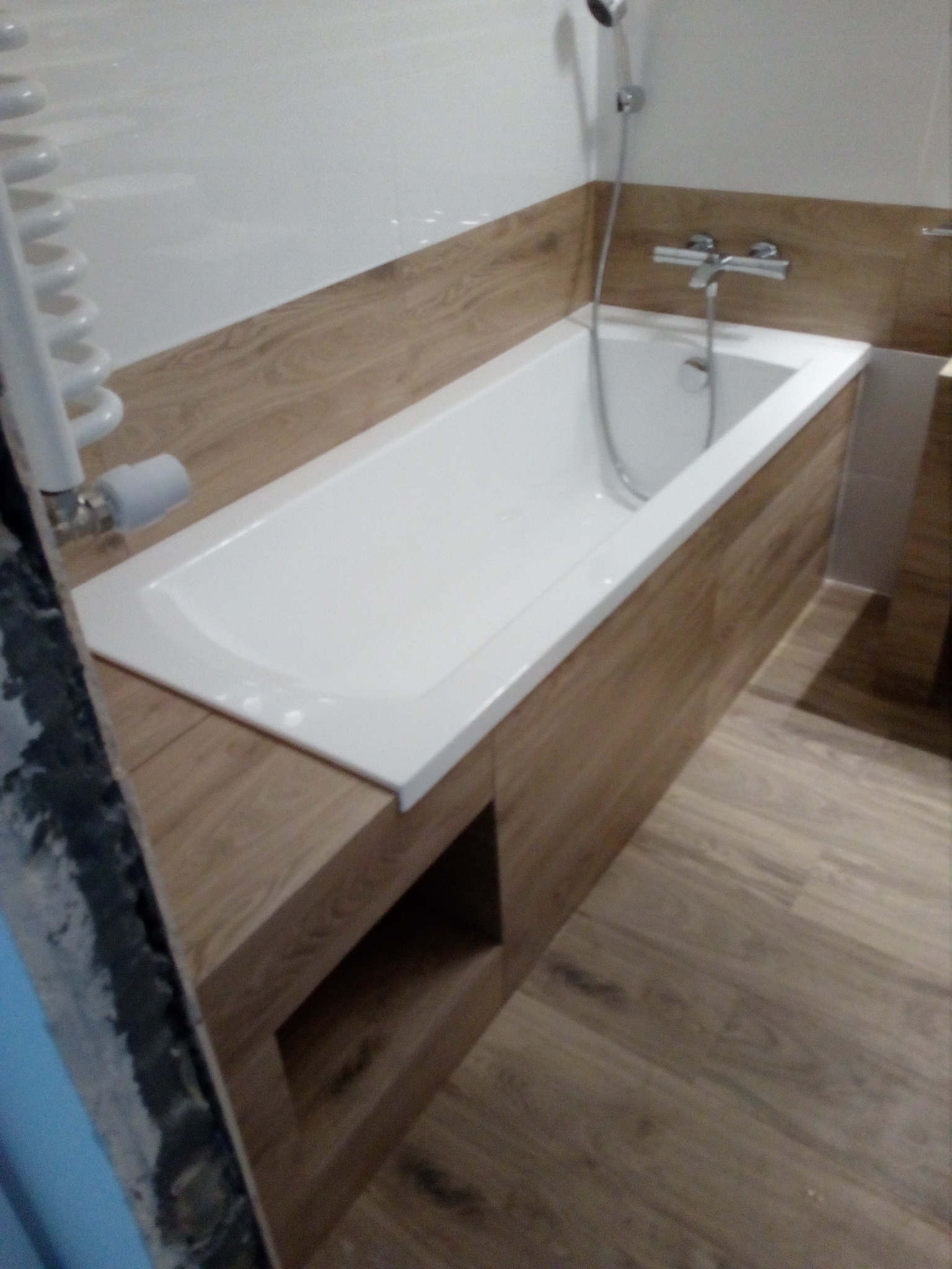 Biała wanna obudowana panelami imitującymi drewno, z widoczną armaturą i grzejnikiem drabinkowym, w trakcie prac wykończeniowych, z fragmentem nieotynkowanej ściany w lewym dolnym rogu.