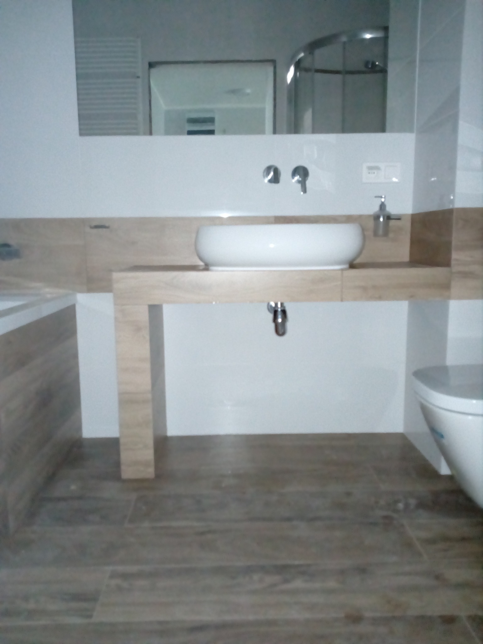 Nowoczesna łazienka z umywalką nablatową na drewnopodobnym blacie, widoczna armatura ścienna, lustro i kabina prysznicowa w tle, płytki podłogowe imitujące drewno.