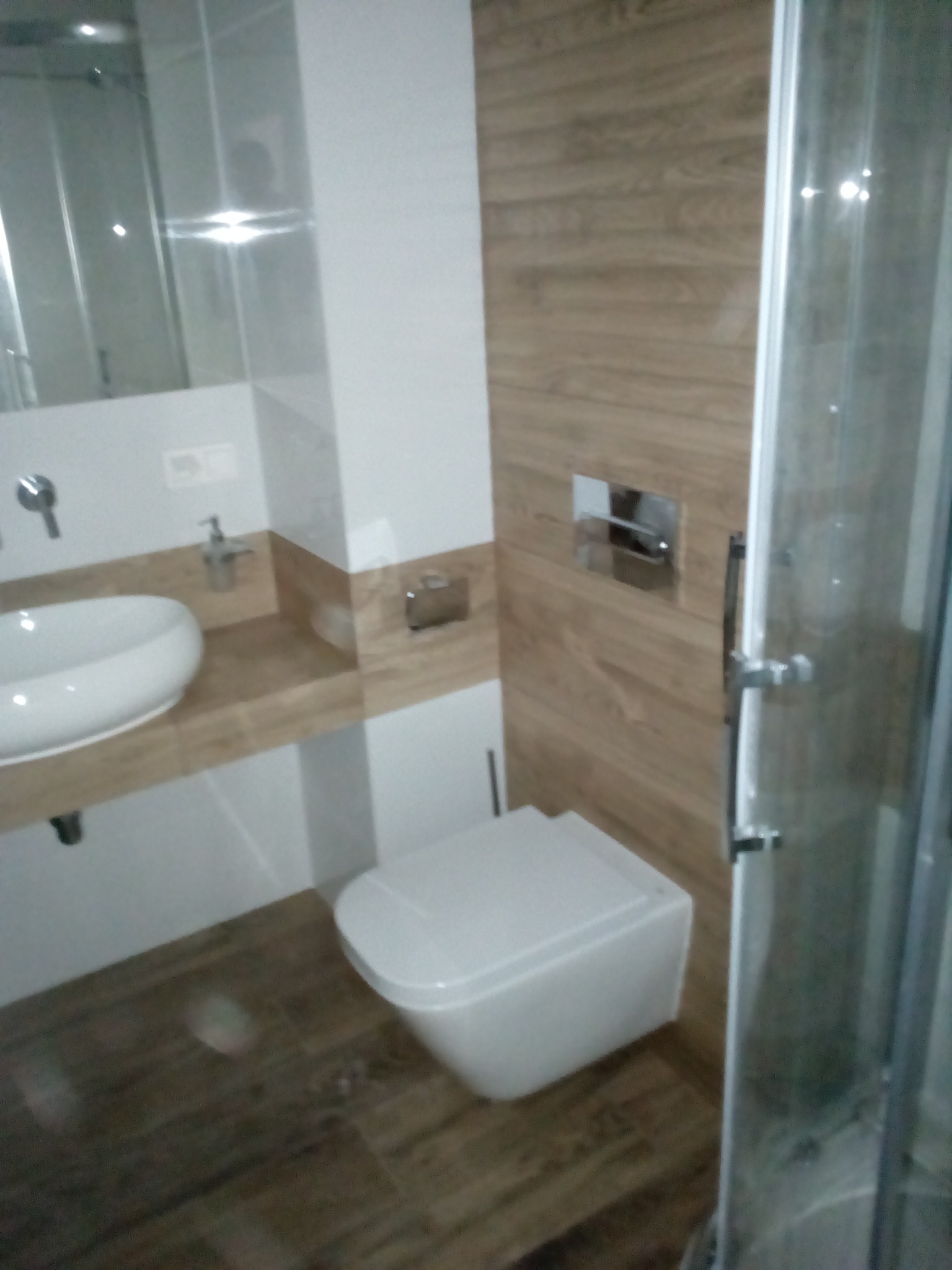 Nowoczesna łazienka z drewnopodobnymi płytkami na ścianie i podłodze, owalna umywalka nablatowa, podwieszana toaleta i częściowo widoczna kabina prysznicowa.