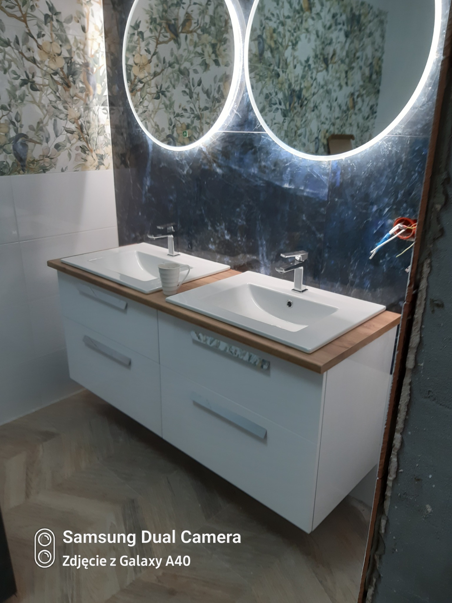 Nowoczesna łazienka z podwójną umywalką nablatową na białej szafce z drewnianym blatem, okrągłe lustra z podświetleniem LED, płytki z motywem roślinnym i marmurowym wzorem, widoczne instalacje...