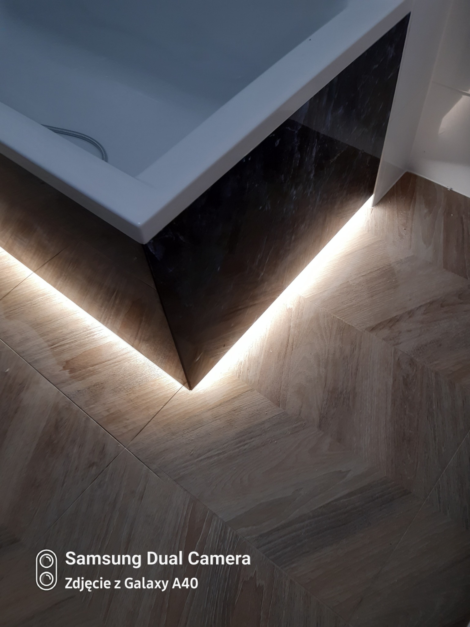 Detal wykończenia łazienki: Podświetlona listwa LED pod wanną z czarną okładziną i parkietem ułożonym we wzór jodełki.