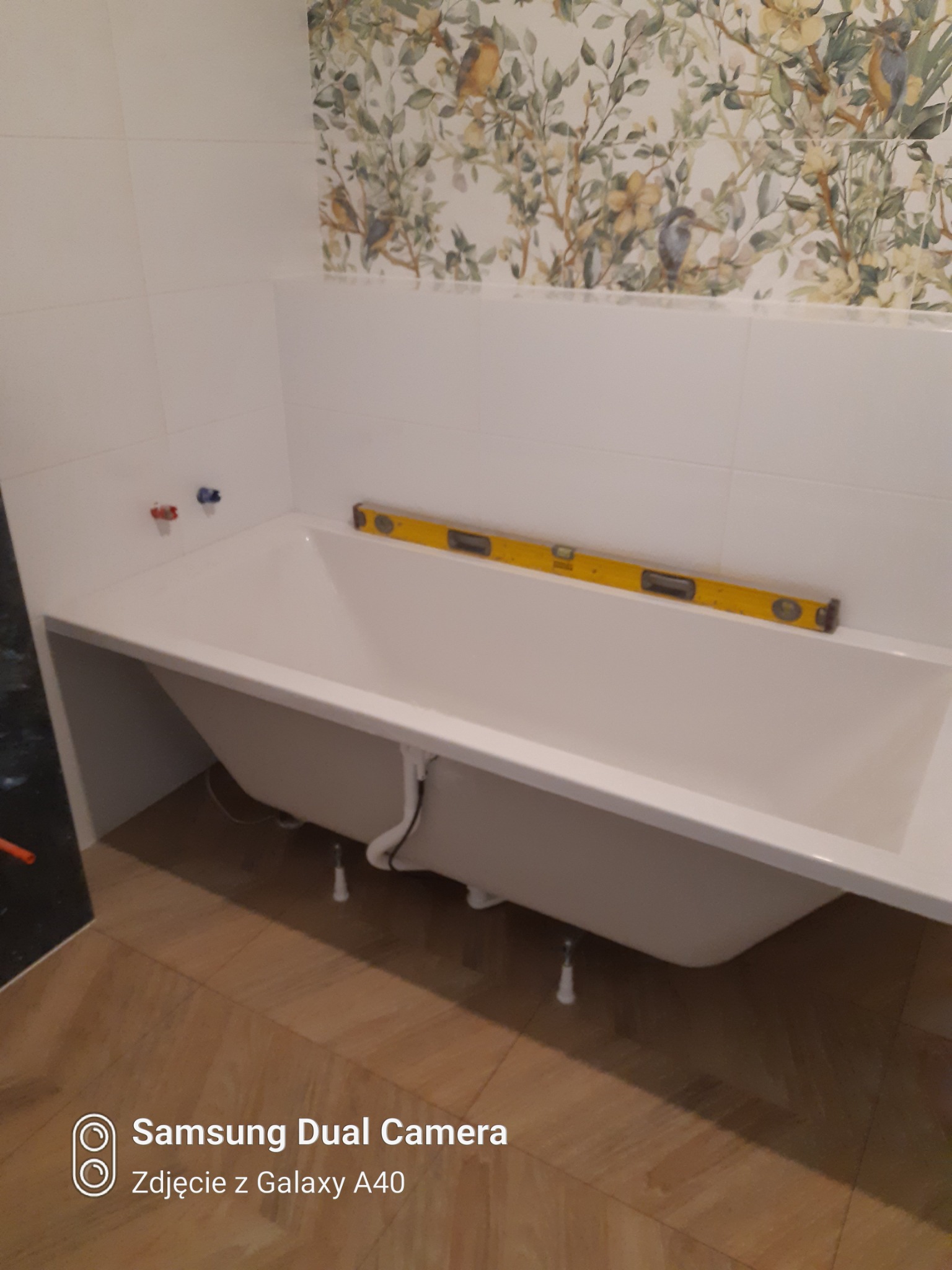 Biała wanna obudowana płytkami, z poziomicą na krawędzi, na tle ściany z motywem roślinnym i ptaków.