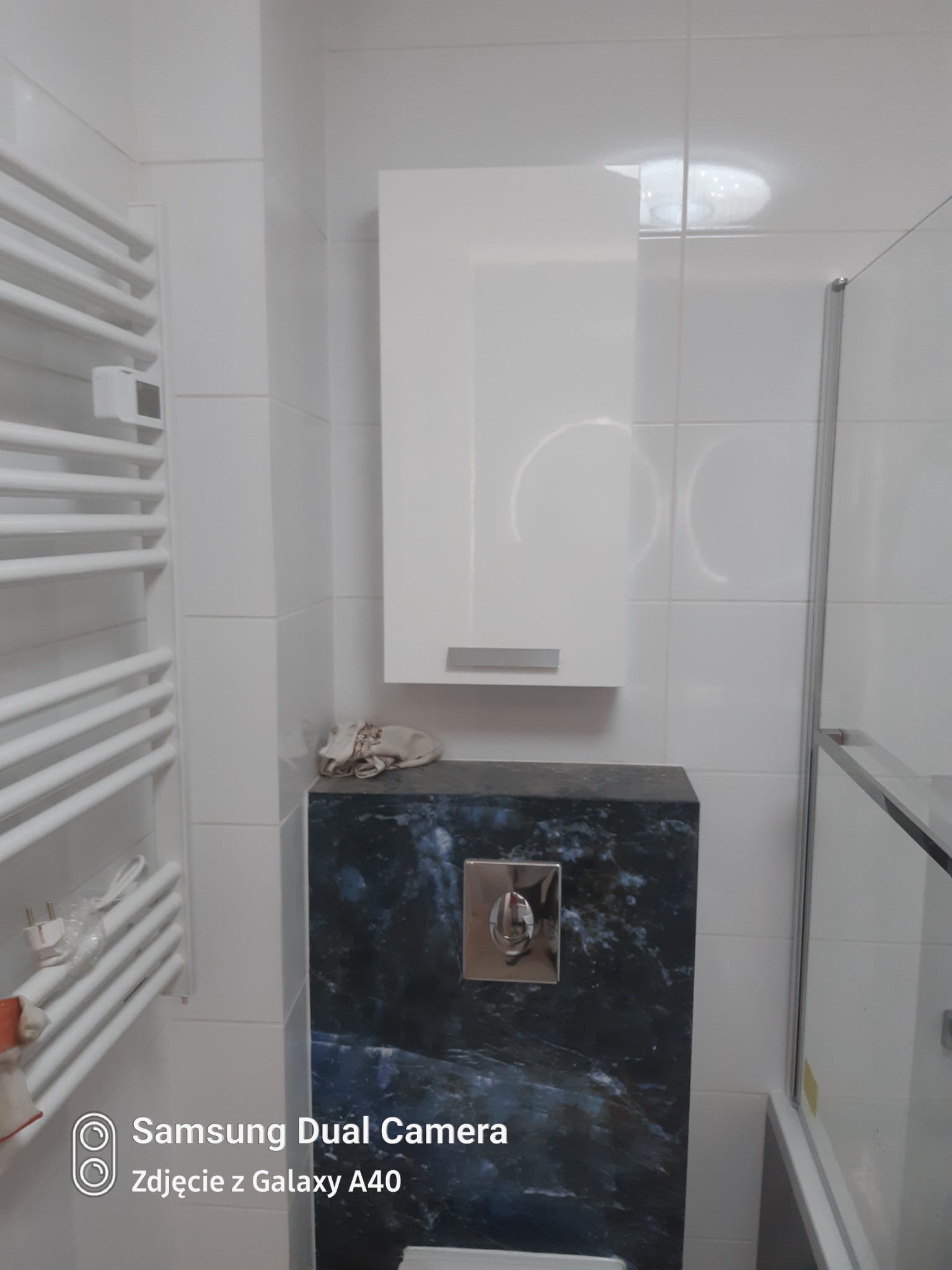 Biała łazienka z grzejnikiem drabinkowym, zabudową toalety w ciemnym marmurze i białą szafką wiszącą, widoczna częściowo kabina prysznicowa.