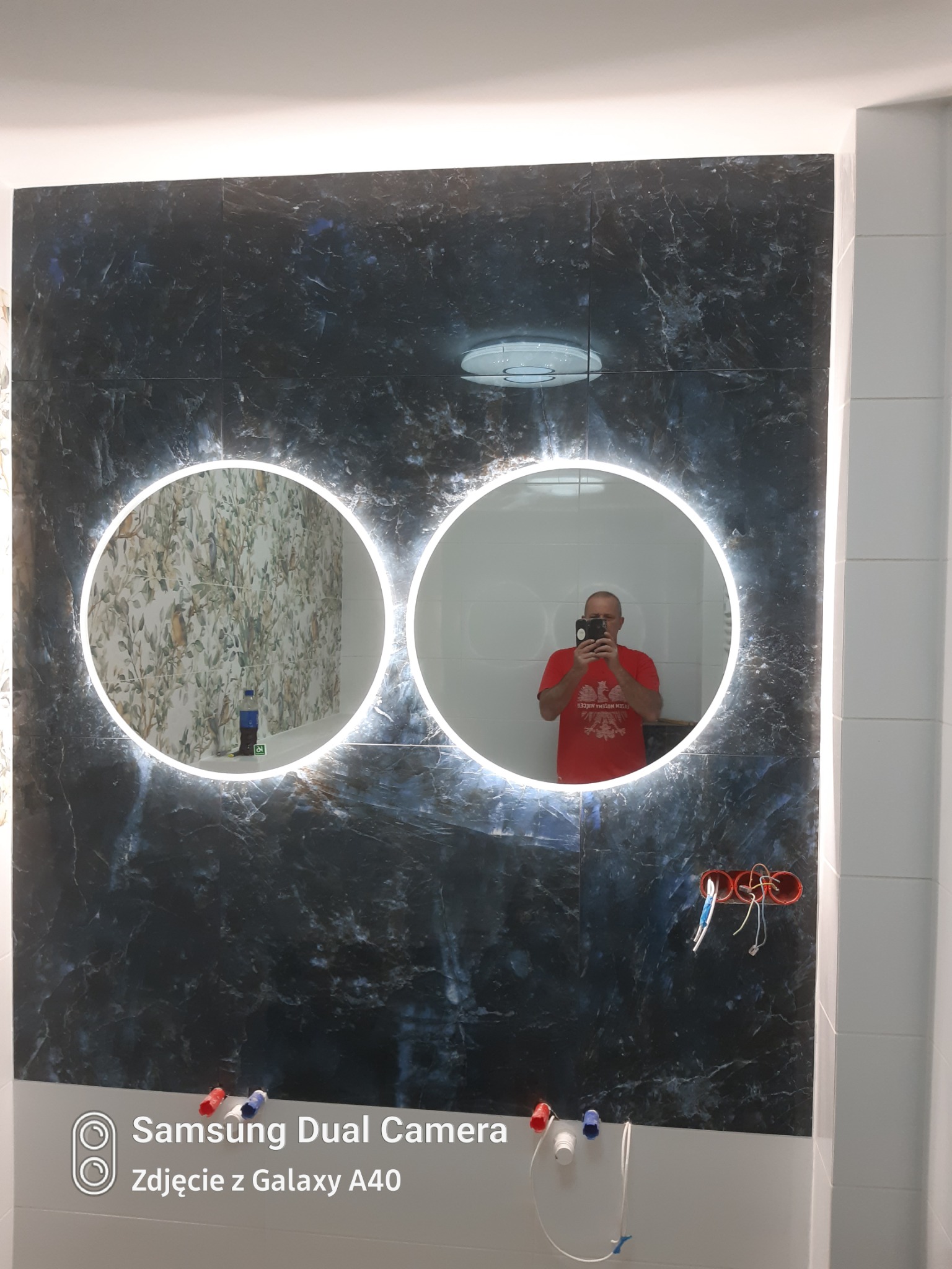 Łazienka z dwoma okrągłymi lustrami z podświetleniem LED na ścianie z płytek imitujących ciemny marmur. Widoczne niezakończone podłączenia elektryczne i hydrauliczne.