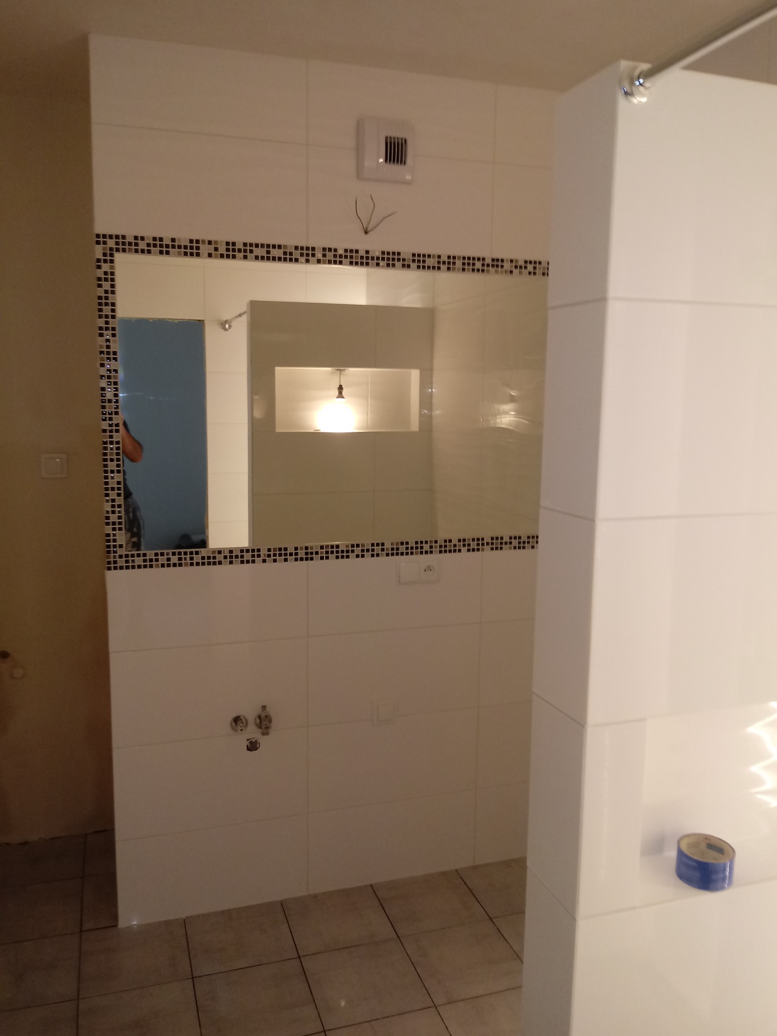 Łazienka w trakcie remontu w Bydgoszczy: widoczne białe płytki z dekoracyjną czarną mozaiką wokół lustra, niezamaskowane przewody wentylacyjne i fragment kolumny z niebieską taśmą malarską.