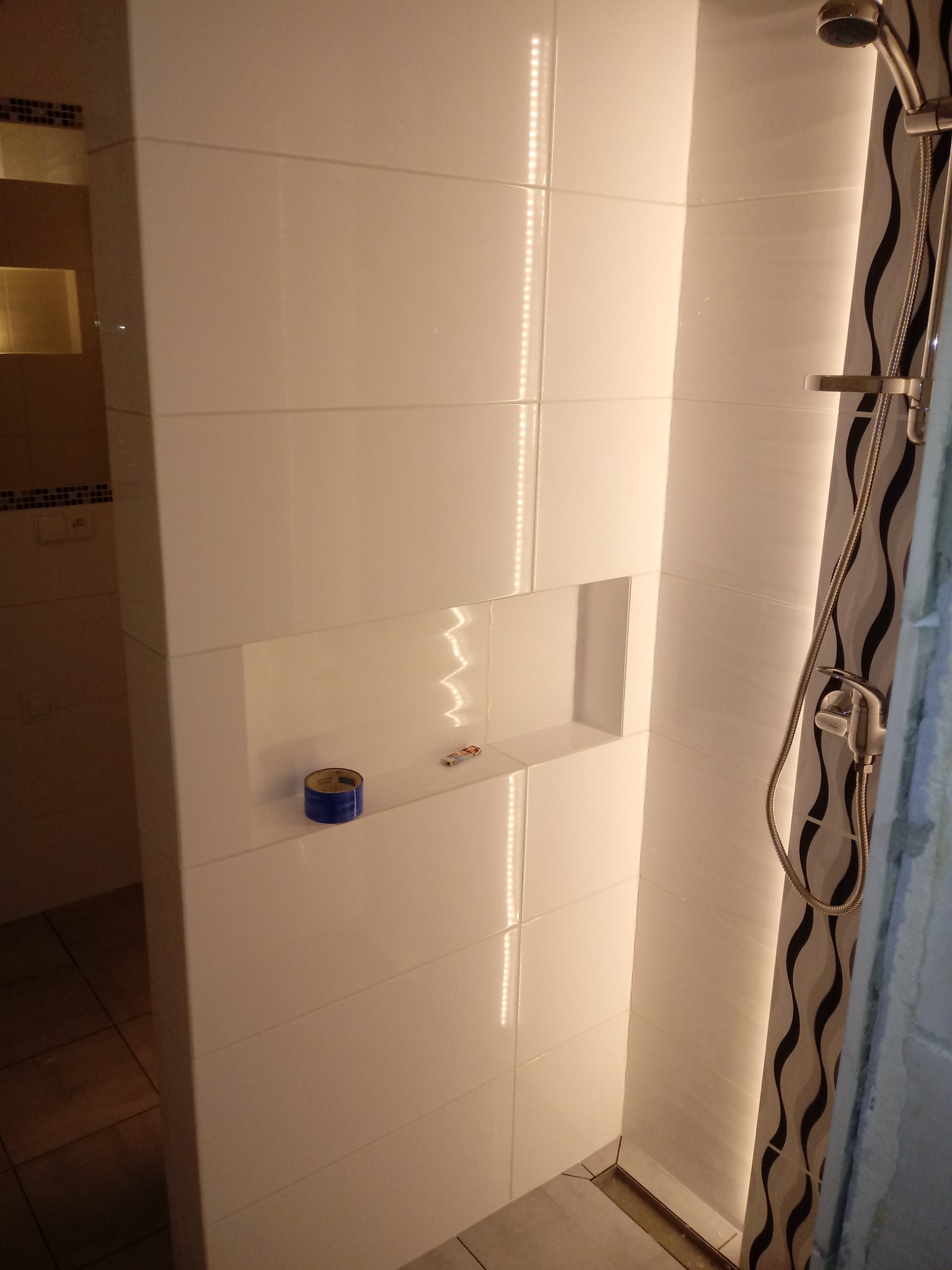 Wykończona kabina prysznicowa z białymi płytkami, wnęką z niebieską taśmą malarską i dekoracyjną listwą z czarnymi falami, oświetlona ledowym paskiem.