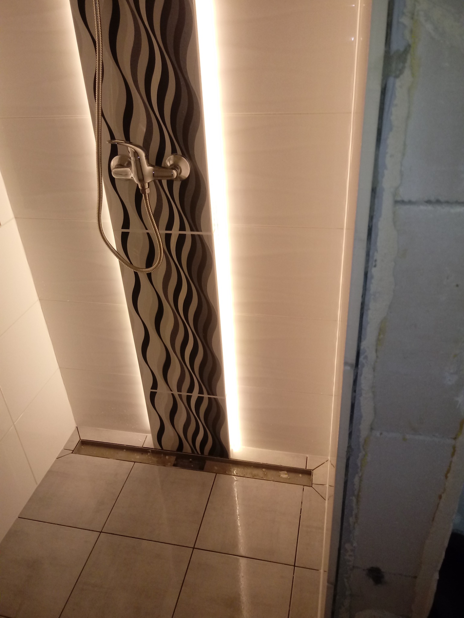 Wykończona kabina prysznicowa z odpływem liniowym, panel dekoracyjny z falistym wzorem podświetlony pionowym paskiem LED, widoczny fragment surowej ściany z betonu komórkowego.