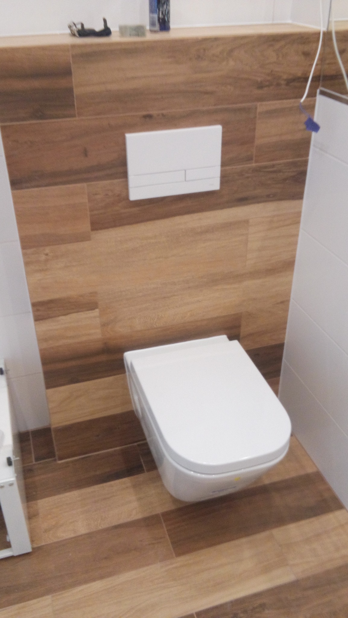 Nowoczesna toaleta podwieszana z białą armaturą i przyciskiem spłukującym na ścianie wyłożonej brązowymi płytkami imitującymi drewno, z fragmentem białej ściany po prawej stronie.