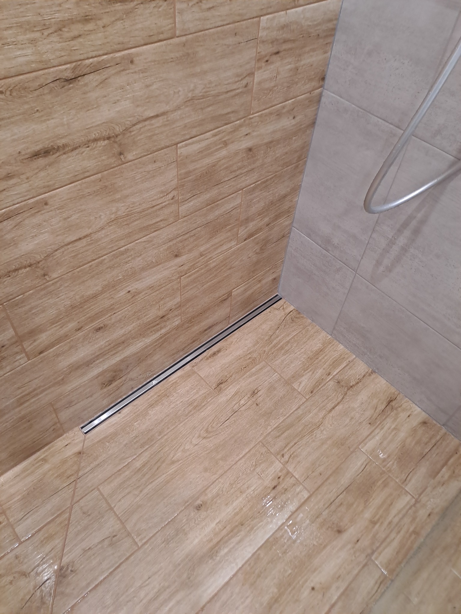 Ukończona łazienka z drewnopodobnymi płytkami na ścianach i podłodze, widoczny odpływ liniowy i fragment kabiny prysznicowej z szarymi płytkami.