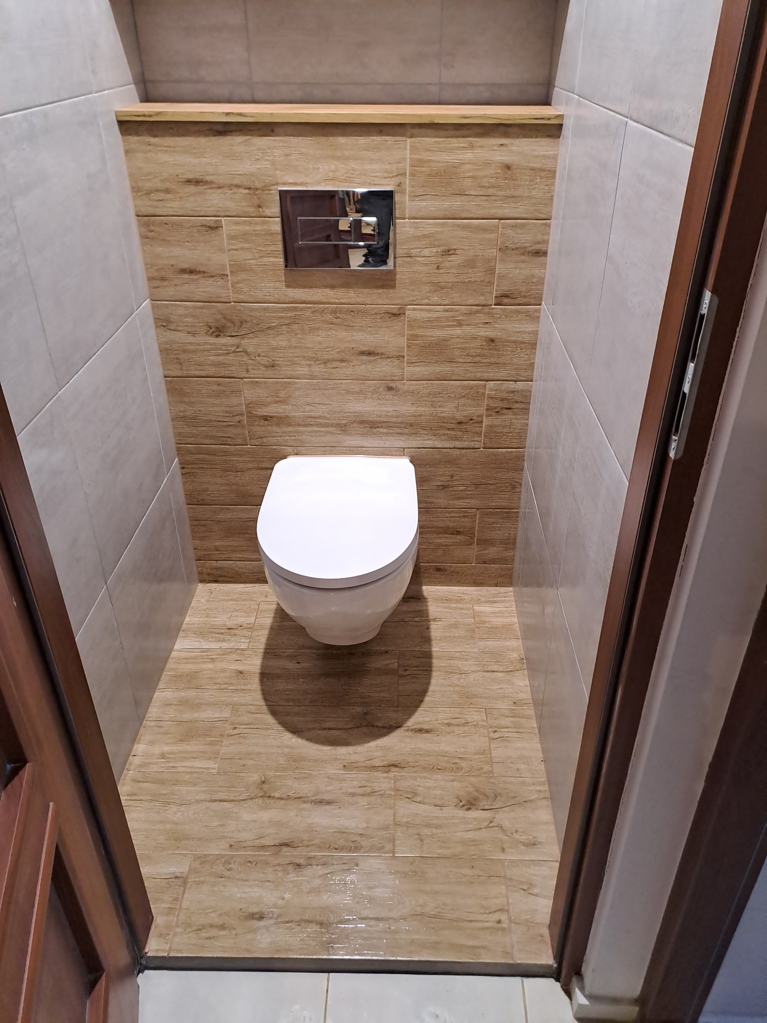 Nowoczesna toaleta w małej łazience wykończonej płytkami imitującymi drewno, z podtynkową spłuczką i widoczną częścią brązowej ościeżnicy.