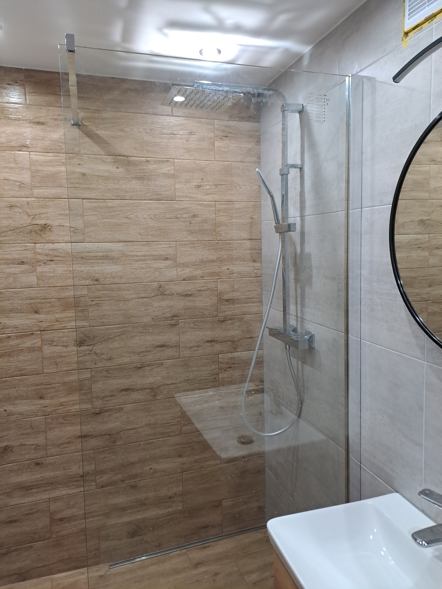 Nowoczesna kabina prysznicowa walk-in z drewnopodobnymi płytkami na ścianach, deszczownicą i szklaną ścianką, widoczna umywalka nablatowa.