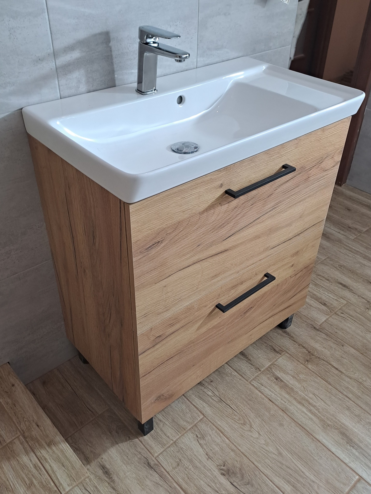 Nowoczesna szafka łazienkowa z umywalką nablatową i chromowaną baterią, fronty w dekorze jasnego drewna z czarnymi uchwytami, na tle szarych płytek ściennych i podłogowych imitujących drewno.