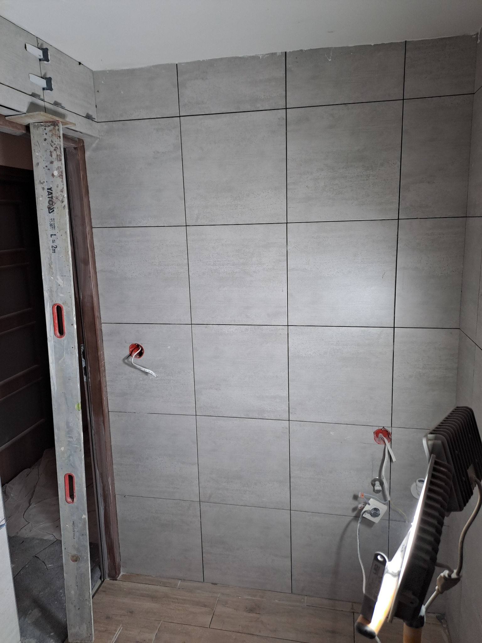 Ściana w łazience wyłożona szarymi płytkami ceramicznymi o fakturze kamienia, widoczne puszki elektryczne bez osprzętu, poziomica oparta o futrynę drzwi, lampa budowlana oświetlająca fragment ściany.