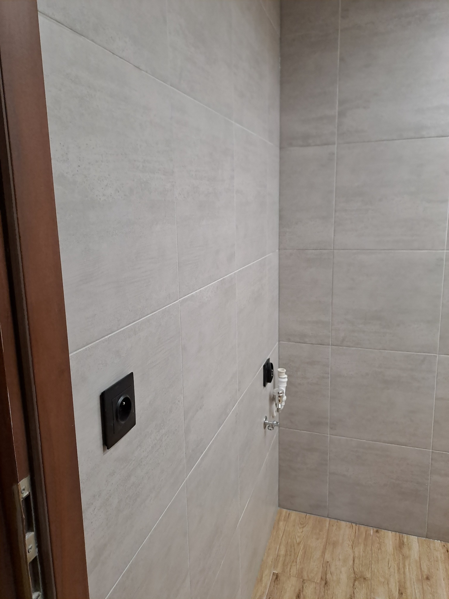 Ściana wyłożona jasnymi, kwadratowymi płytkami ceramicznymi z widocznym gniazdkiem elektrycznym w czarnej obudowie i zaworem wodnym z białym wężykiem, przy drewnianej ościeżnicy drzwiowej,...