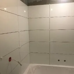 Świeżo ułożone, błyszczące, białe płytki ceramiczne na ścianach łazienki z widocznymi krzyżykami dystansowymi, surowy beton na górze ściany i wystający przewód elektryczny ze ściany.