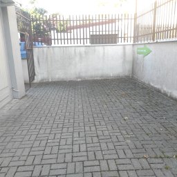 MoMa Maciej Strupiński - Szara kostka brukowa ułożona w regularny wzór, z widocznymi spoinami, niewielkimi chwastami i liśćmi, przylegająca do szarego, lekko zabrudzonego muru z zieloną strzałką informacyjną i metalowym...