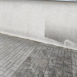 MoMa Maciej Strupiński - Świeżo ułożona, mokra kostka brukowa w odcieniach szarości przylegająca do ściany budynku z widocznymi zaciekami i zabrudzeniami.