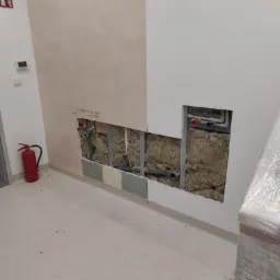 Ściana w trakcie remontu z odsłoniętą instalacją hydrauliczną i izolacją, widoczny fragment pomalowanej ściany w dwóch odcieniach beżu, gaśnica w rogu pomieszczenia.