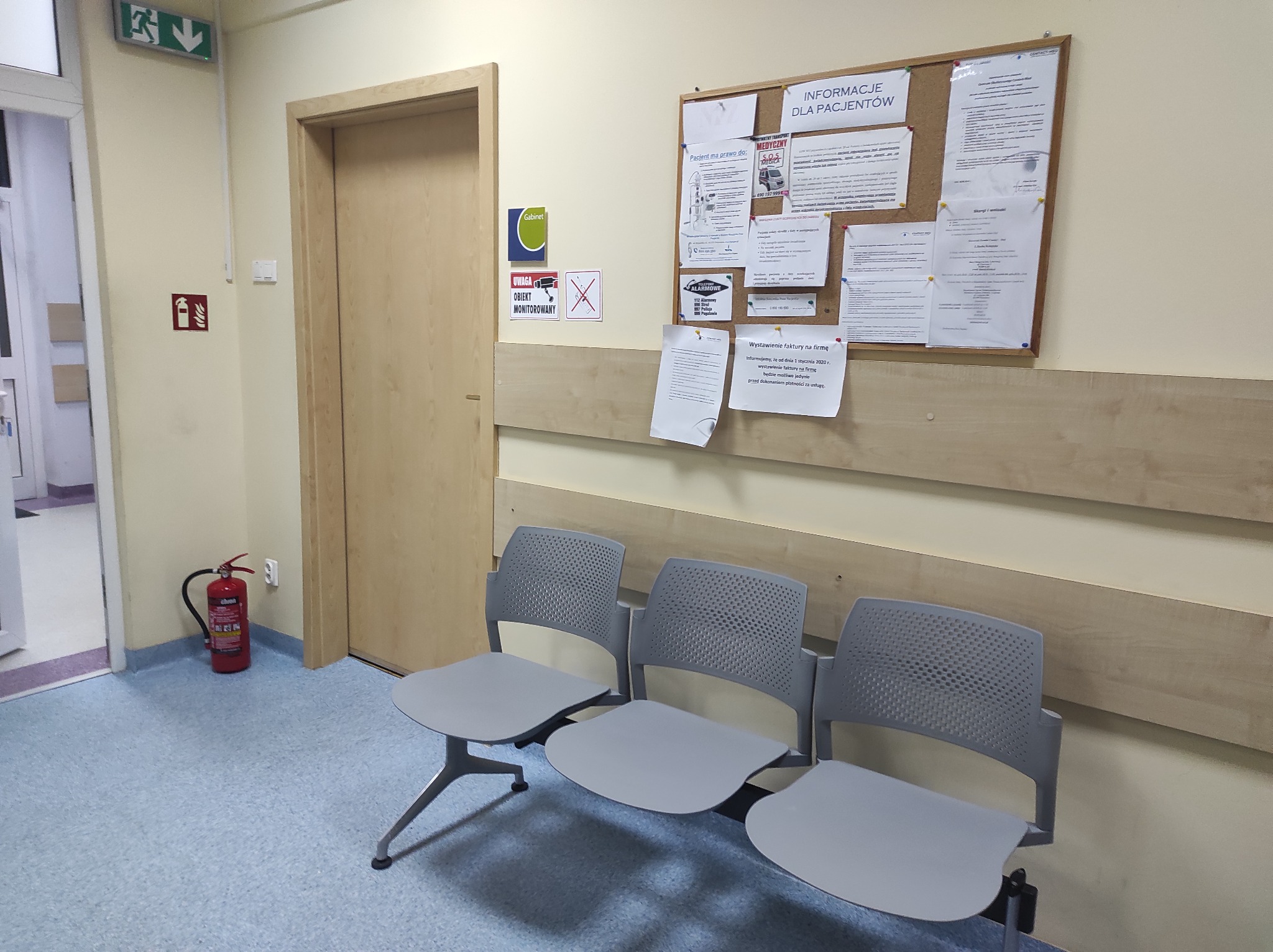 Poczekalnia w przychodni: trzy szare krzesła z perforowanymi oparciami ustawione pod ścianą z tablicą informacyjną i wywieszkami, widoczna gaśnica i znak ewakuacyjny.