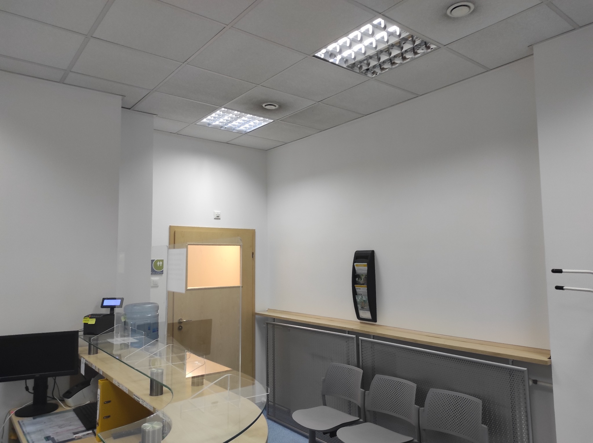 Jasne, minimalistyczne wnętrze biurowe z recepcją, szklaną osłoną, szarymi krzesłami i oświetleniem sufitowym.