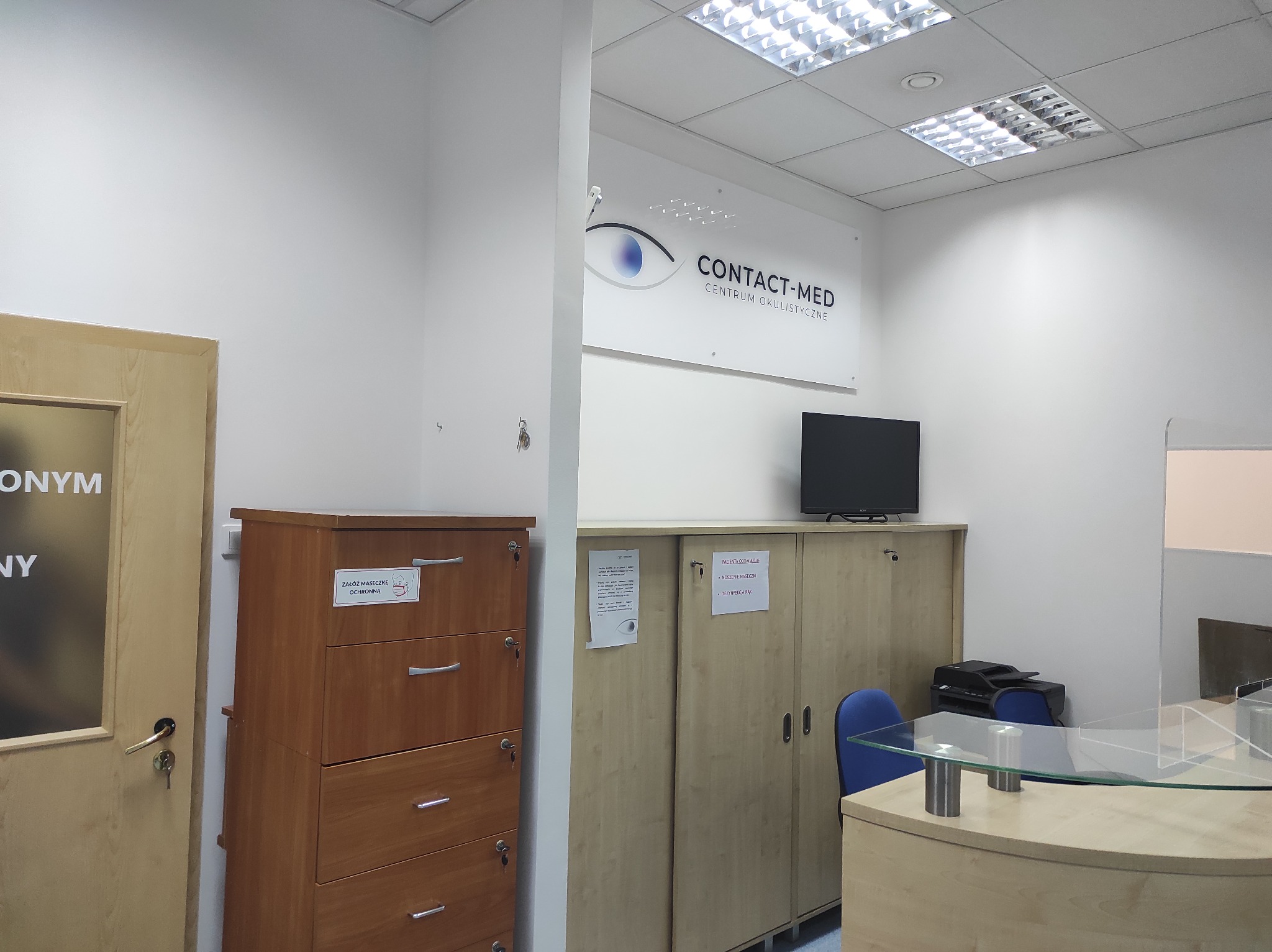 Wnętrze biura centrum okulistycznego z recepcją, szafkami na dokumenty i logo firmy Contact-Med na ścianie.