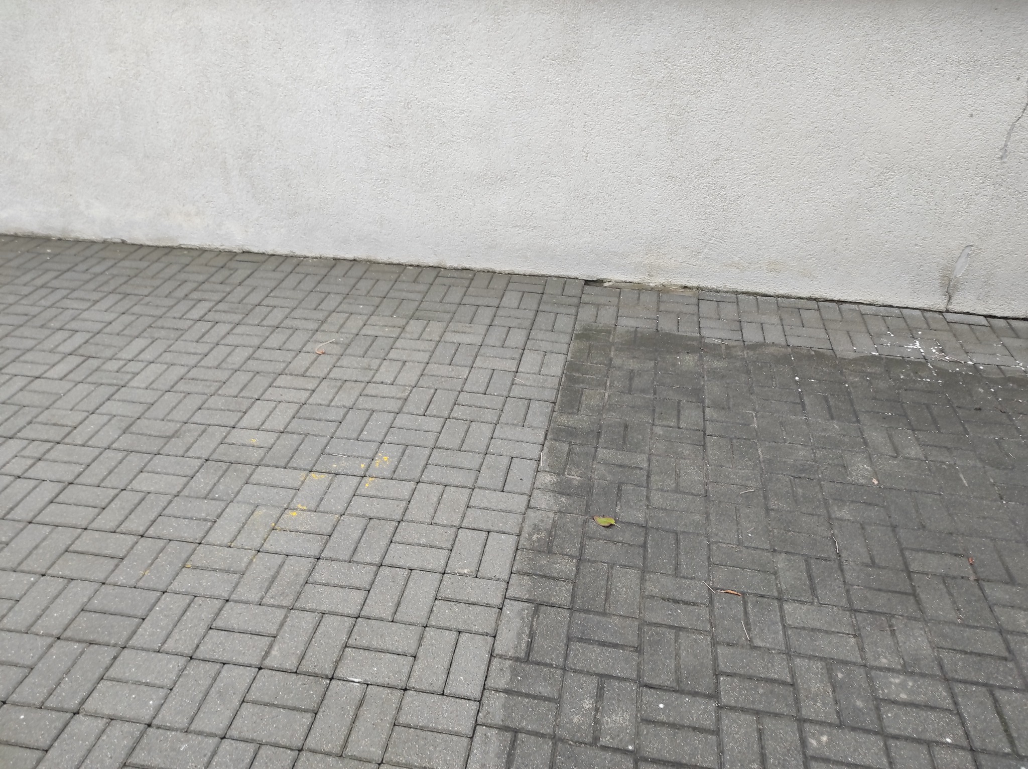 Świeżo ułożona, szara kostka brukowa w jodełkę, częściowo mokra, przylegająca do szarej, chropowatej ściany budynku.