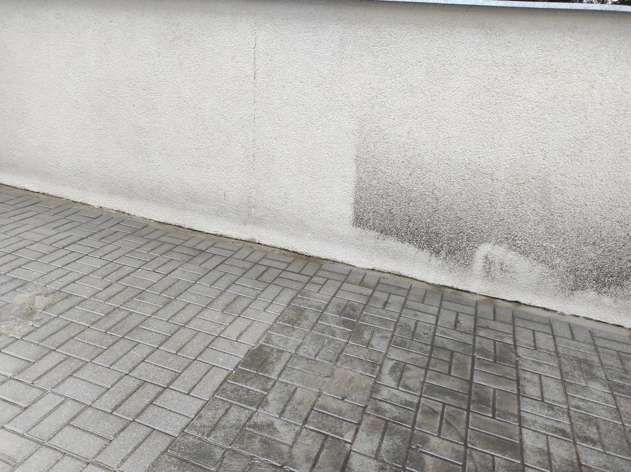 Świeżo ułożona, mokra kostka brukowa w odcieniach szarości przylegająca do ściany budynku z widocznymi zaciekami i zabrudzeniami.