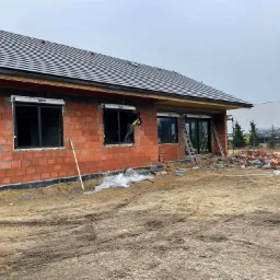 Budowa domu jednorodzinnego z czerwonej cegły, widoczny murarz montujący roletę zewnętrzną w oknie, teren wokół w stanie surowym.