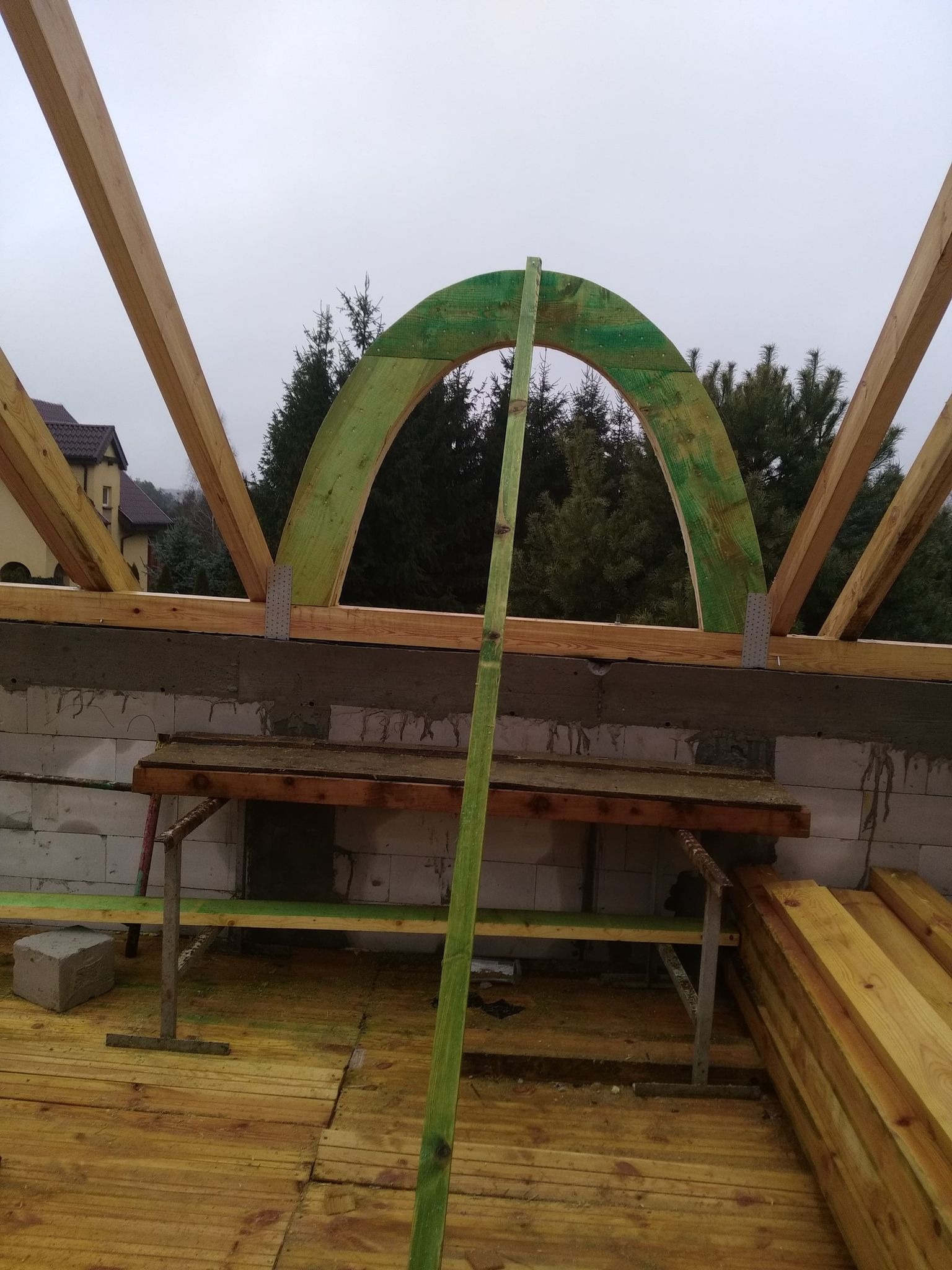 Wnętrze w trakcie budowy z elementami drewnianymi, w tym zielony łuk okienny, widoczne bloczki betonowe i rusztowanie